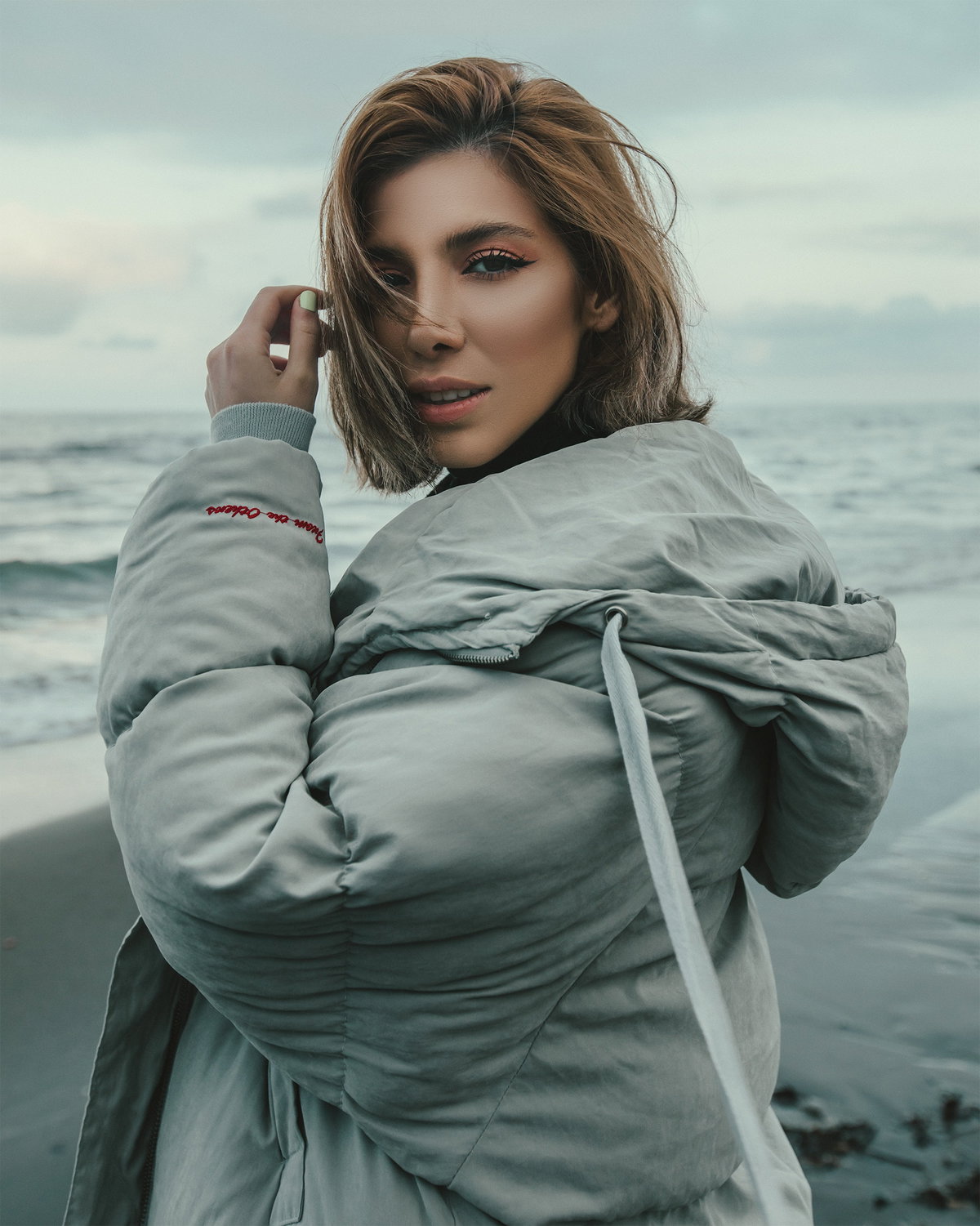 Junge Frau am Strand mit Daunenjacke und Kurzhaarfrisur