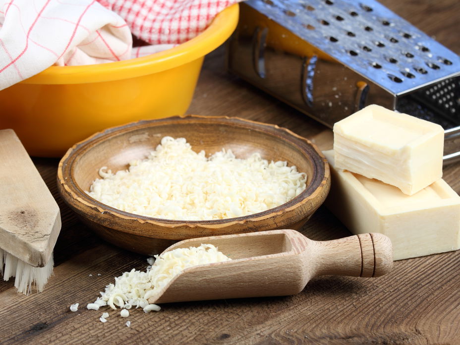 Waschmittel selber machen: Kernseife (gerieben) in einer Holzschüssel neben Seife, einer Reibe, einer Bürste und einer Schüssel auf einem Holztisch
