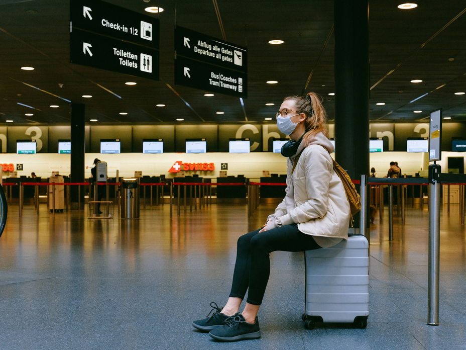 Frau sitzt am Flughafen mit Maske und Koffer