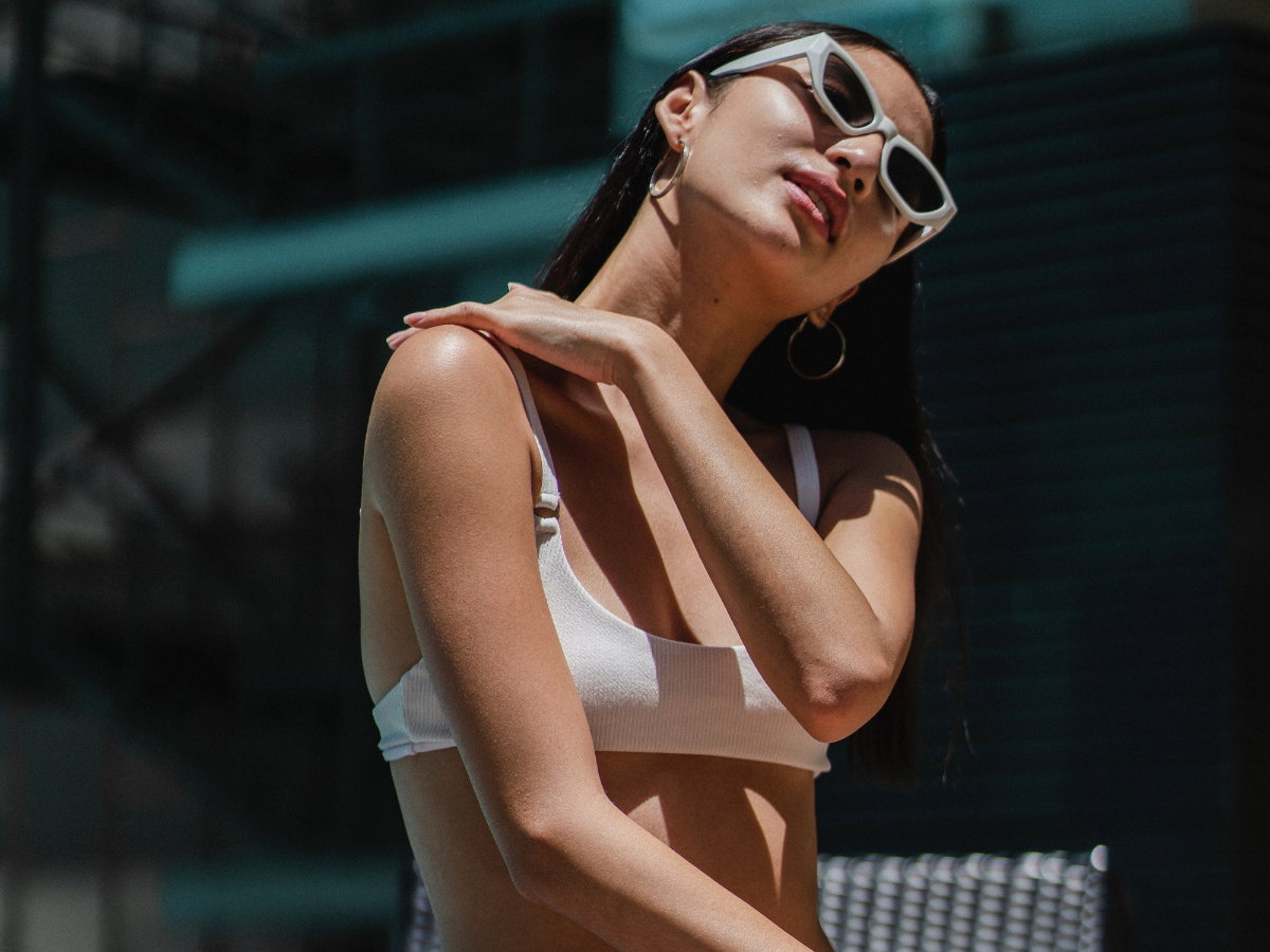 Eine Frau mit Bikinioberteil und Sonnenbrille trägt Sonnenschutz am Hals auf.