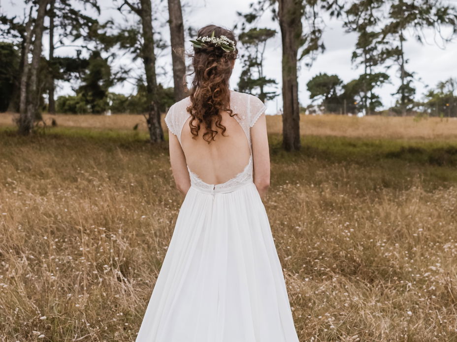 Frau mit Boho Hochzeitsfrisur steht auf einem Feld mit dem Rücken zur Kamera.