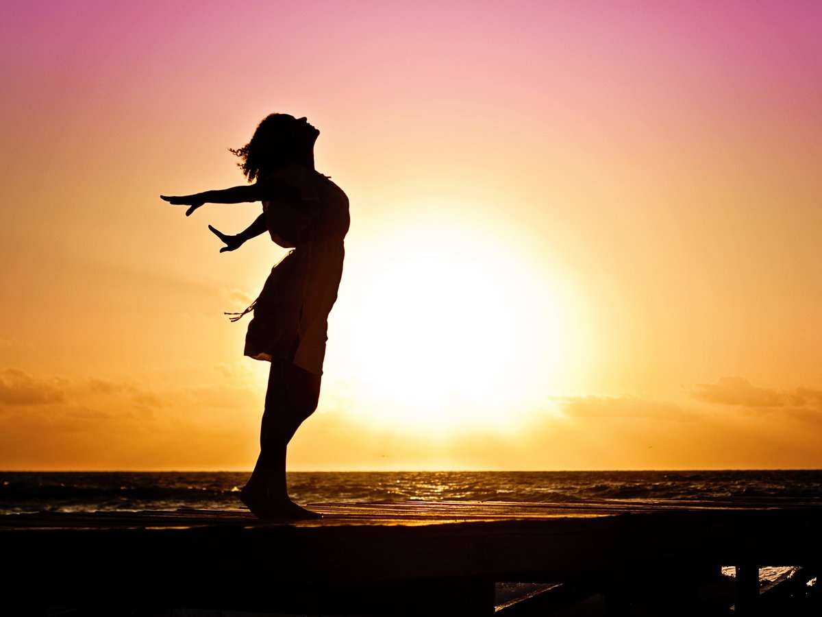Silhouette einer Frau vor einem Sonnenaufgang, die die Arme nach hinten ausstreckt