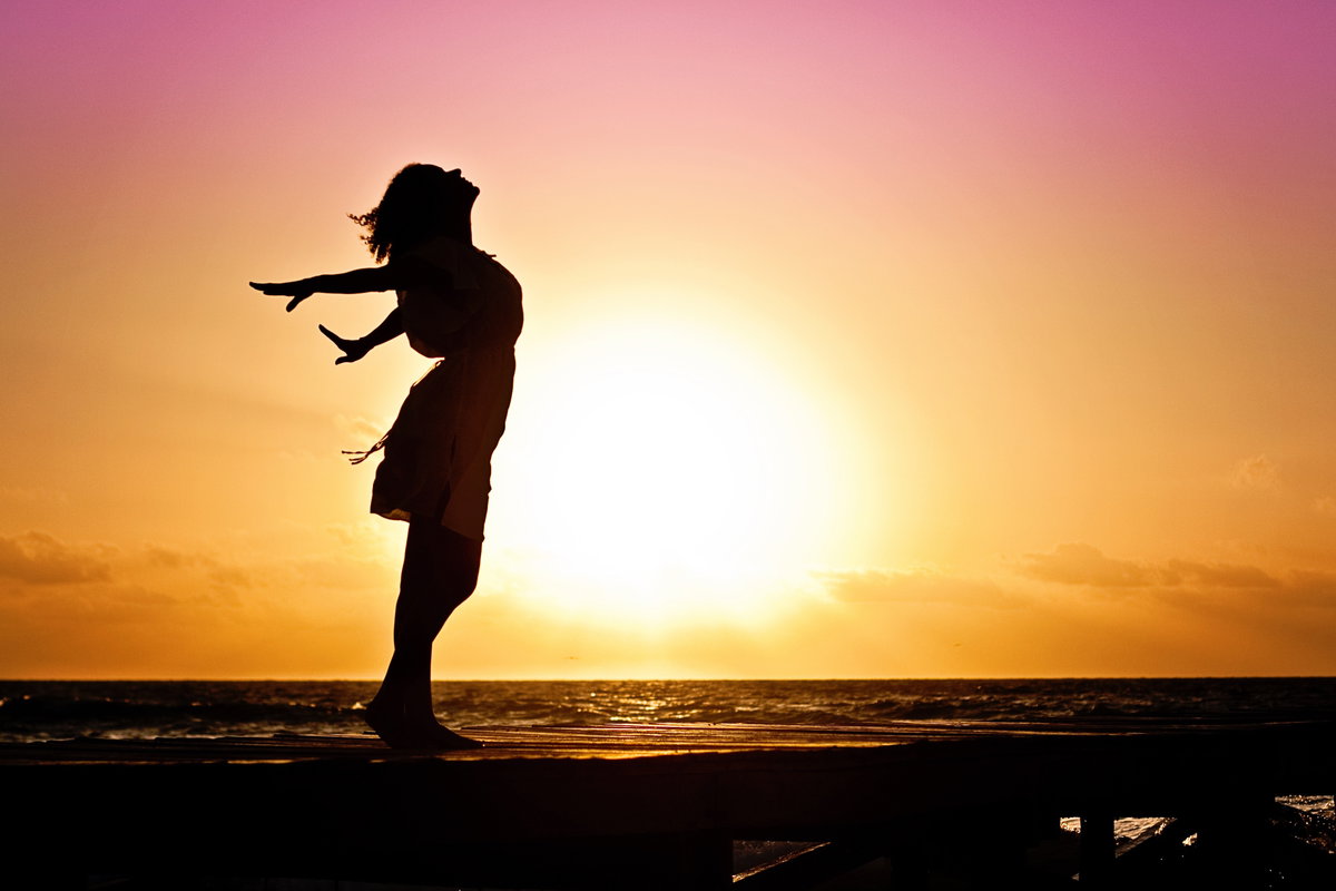 Silhouette einer Frau vor einem Sonnenaufgang, die die Arme nach hinten ausstreckt