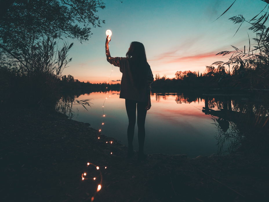 Frau am See hält eine Glühbirne mit einer Lichterkette in der Hand.