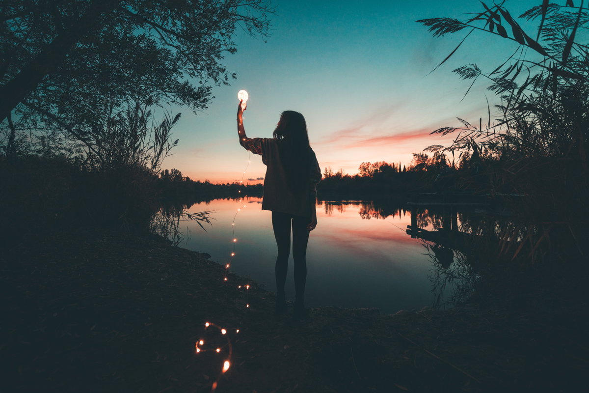 Frau am See hält eine Glühbirne mit einer Lichterkette in der Hand.