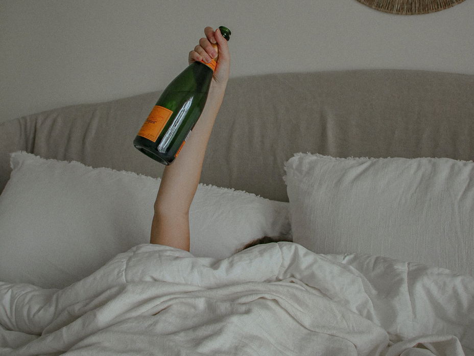 Frau im Bett hält mit einem Arm eine Flasche Alkohol in die Luft