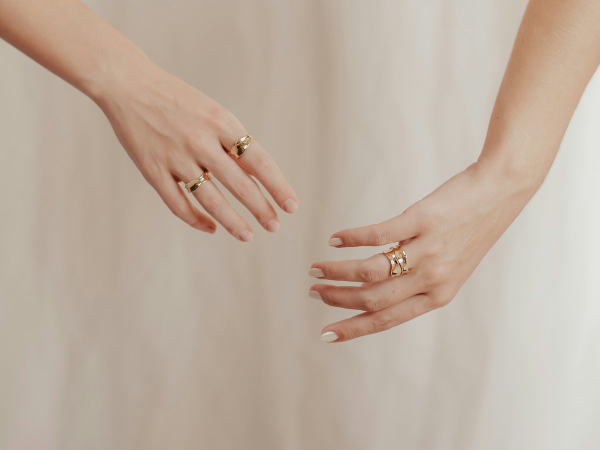 Zwei Hände vor beigem Hintergrund. An beiden Händen sind goldene Ringe an den Händen zu sehen.