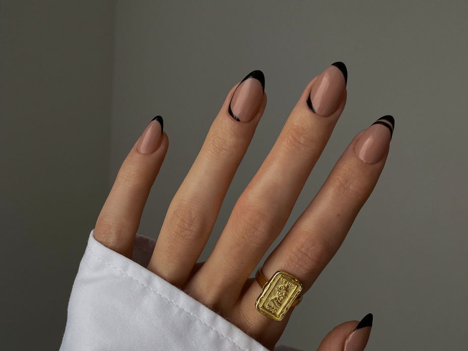 Die Hand einer Frau mit schwarzen schwarzen French Nägeln und einem goldfarbenen Ring am Zeigefinger.