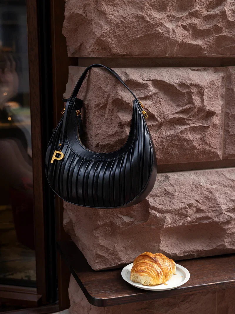 Eine schwarze Schultertasche hängt an einer Wand, darunter steht ein Croissant.