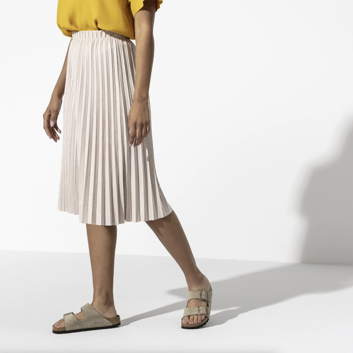 Eine Frau trägt einen Midi-Plisseerock, ein gelbes Oberteil und taupefarbene Birkenstock Sandalen.