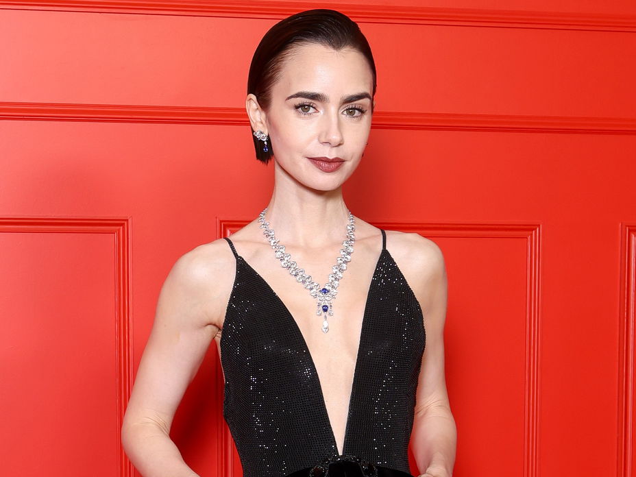 Lily Collins in einem schwarzen Abendkleid mit tiefem V-Ausschnitt vor einer roten Wand.