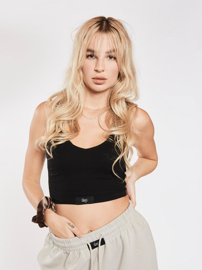 Eine junge Frau mit blonden langen Haaren, einem schwarzen Top und einer grauen Jogginghose.