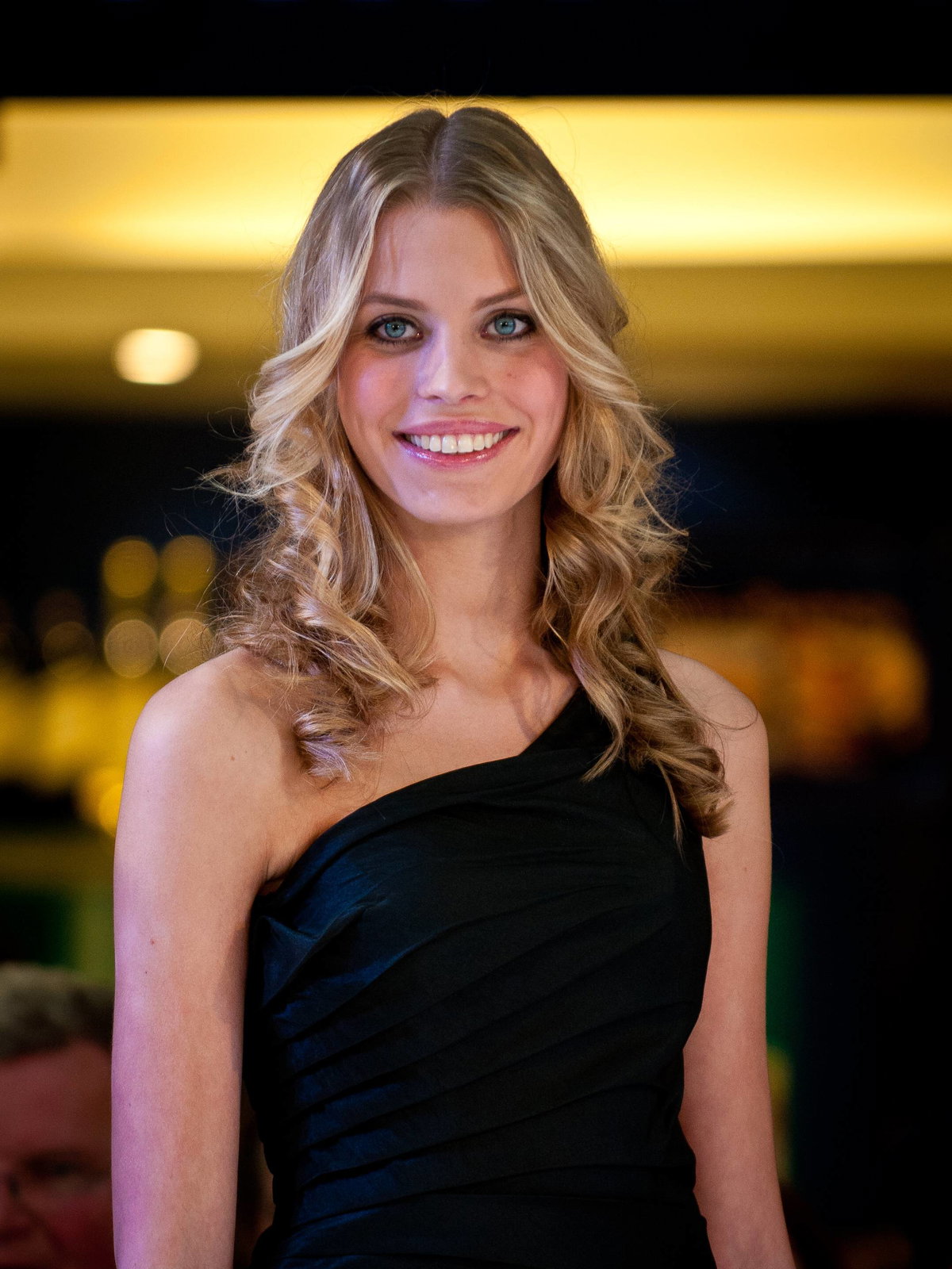 Eine junge Frau mit blonden, lockigen Haaren, großen Augen und einem schwarzen One-Shoulder-Kleid.