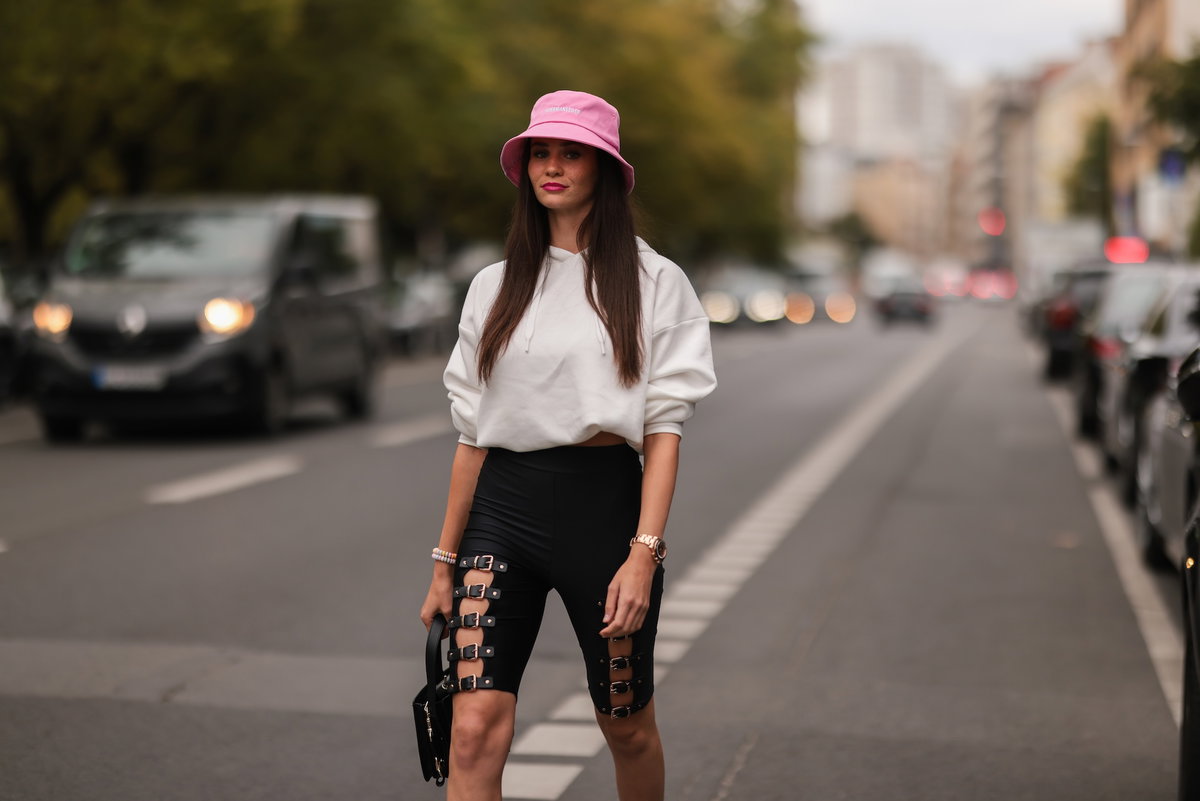 Betty Taube mit einem weißen Sweatshirt, einem pinken Bucket Hat und einer schwarzen Radlerhose mit Schnallen.