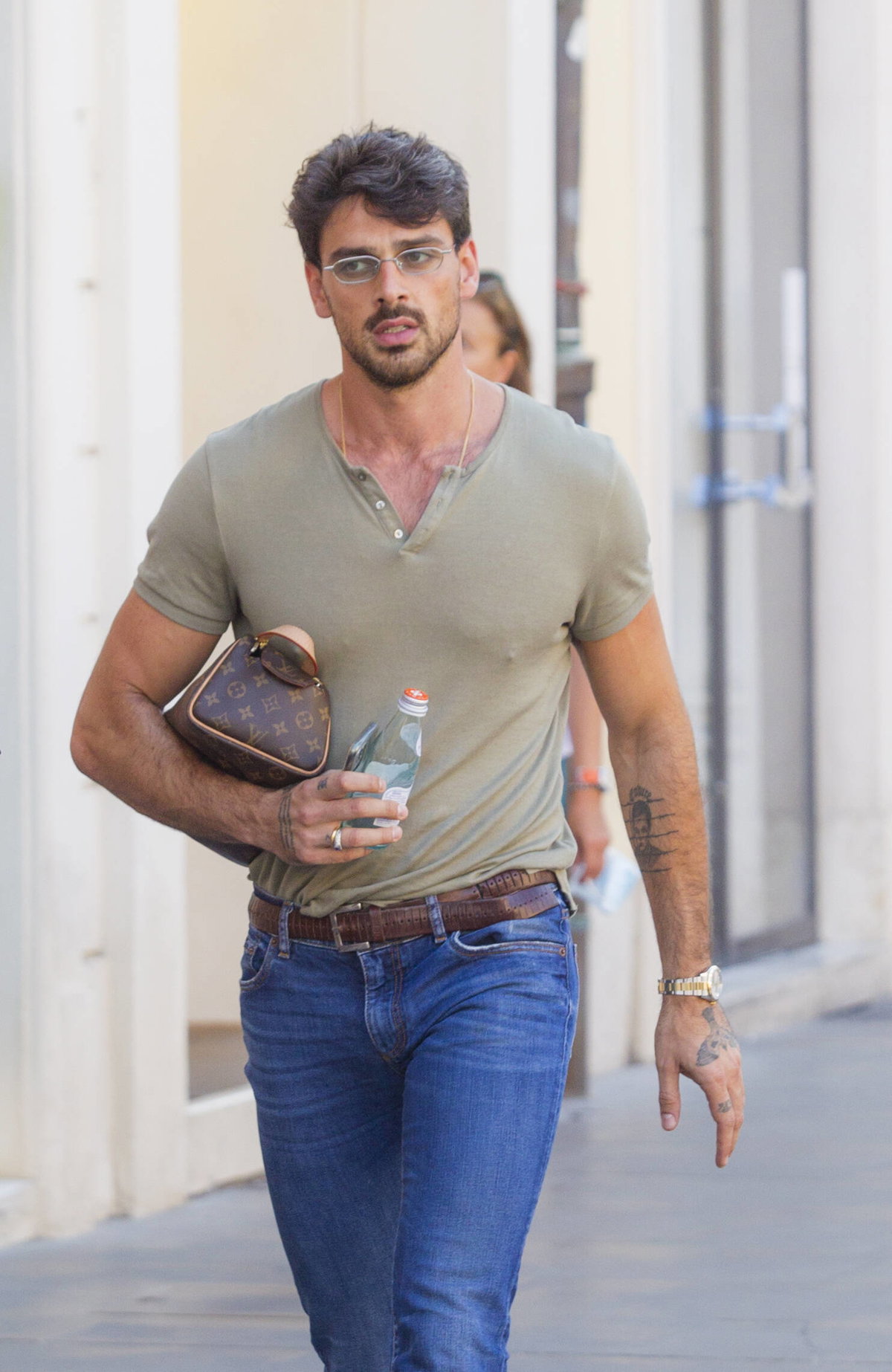 Michele Morrone mit einem khakifarbenen Shirt, einer blauen Jeans, einer Brille und einer Louis Vuitton Tasche auf der Straße unterwegs.