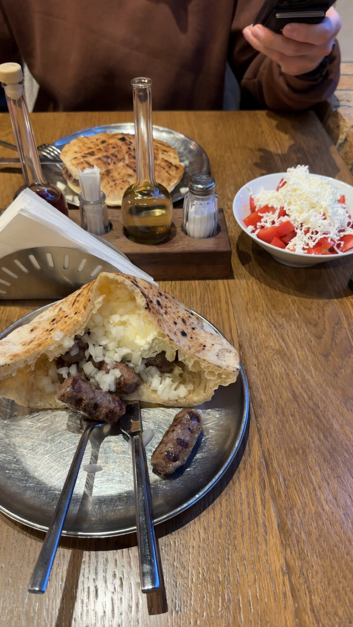 Cevapi im Brötchen mit Zwiebeln auf einem Silberteller auf einem Holztisch mit Salz, Pfeffer, Öl und einem Tomatensalat mit Käse.