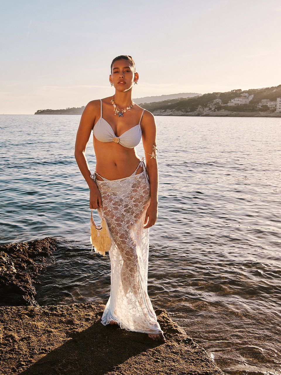 Eine junge Frau steht auf einem Felsen im Wasser und trägt einen weißen Bikini und einen Spitzenrock darüber.