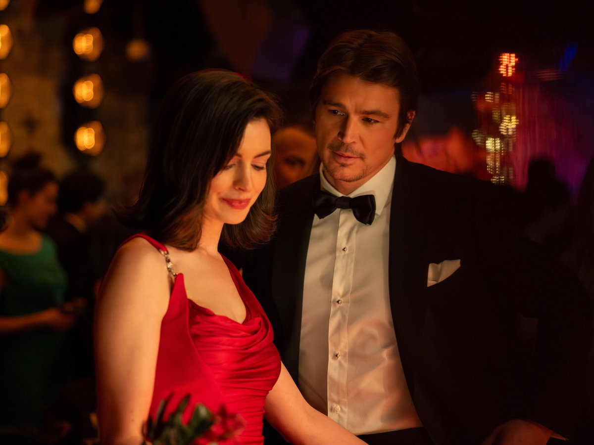 Anne Hathaway steht in einem roten Kleid und schaut nach unten, Josh Hartnett steht hinter ihr und schaut sie an.