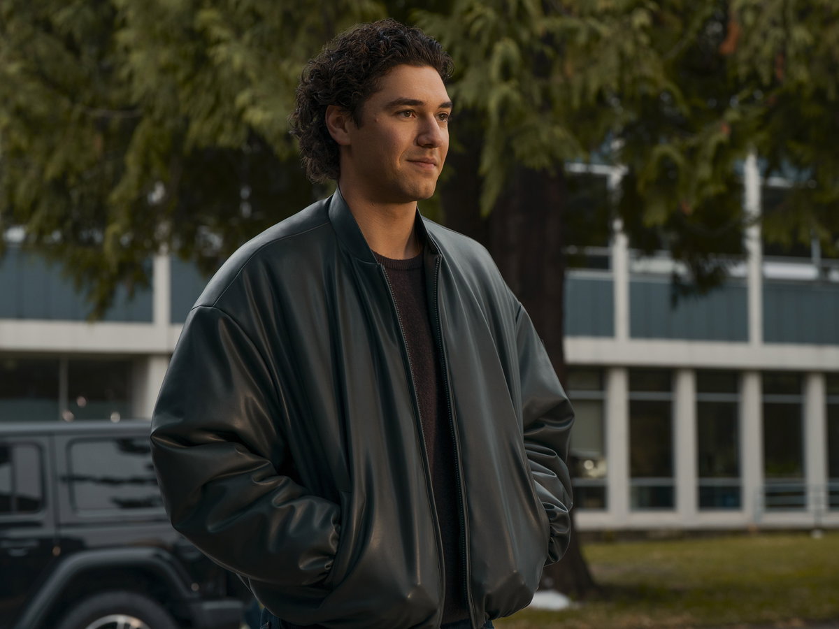 Ein junger Mann mit dunklen Locken in einer Lederjacke steht mit seinen Händen in der Tasche und guckt zur Seite.
