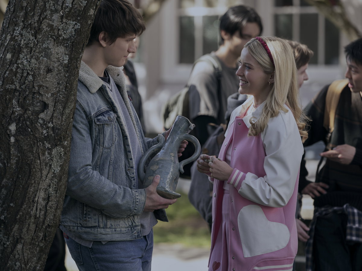 Eine junge Frau mit einer pink-weißen Bomberjacke und blonden Haaren spricht draußen mit einem jungen Mann in einer Jeansjacke, der eine Vase in der Hand hält.