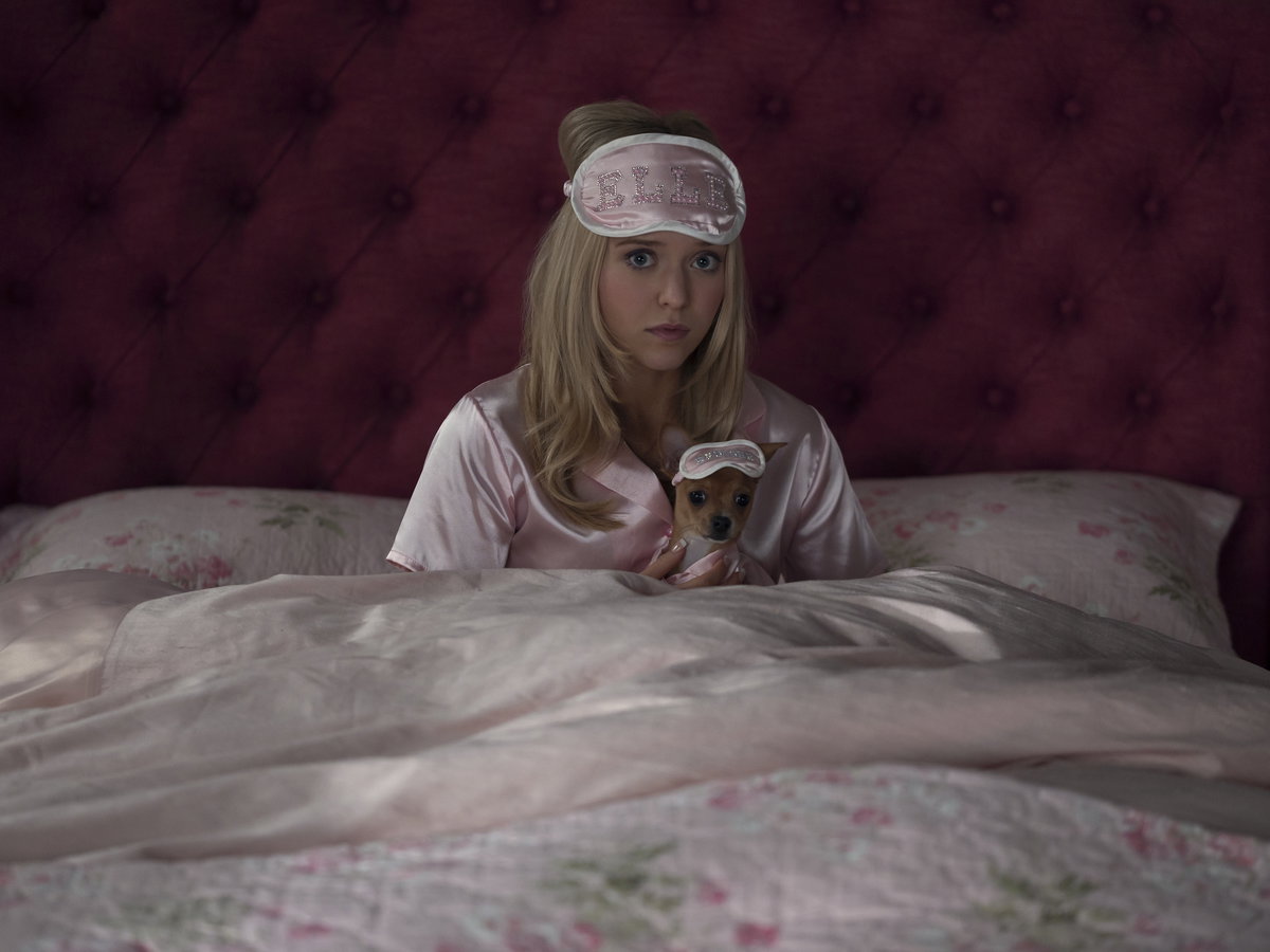 Eine junge Frau mit blonden Haaren sitzt mit einem rosanen Satin-Schlafanzug und Schlafmaske im Bett und hat einen Chihuahua mit Schlafmaske in der Hand.