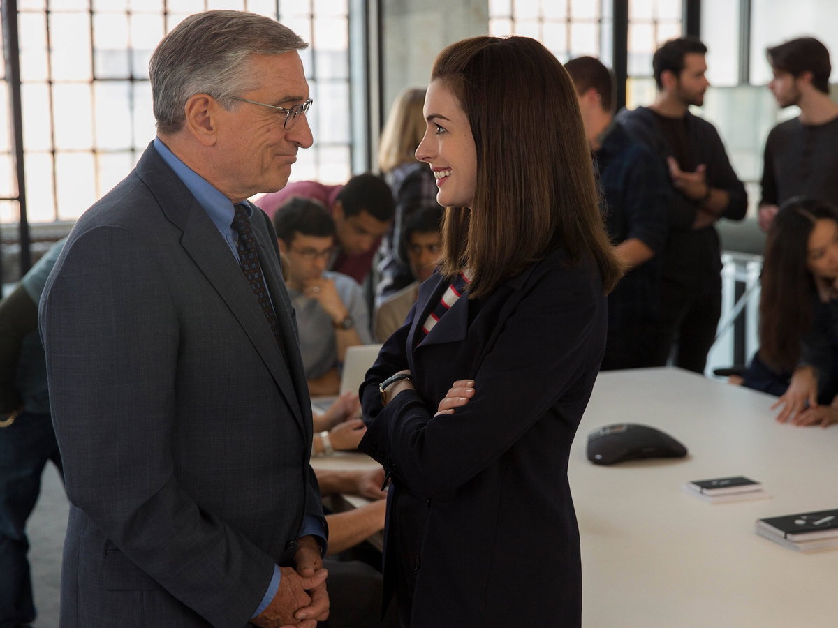 Anne Hathaway und Robert de Niro stehen in einem vollen Büro und lächeln sich an.