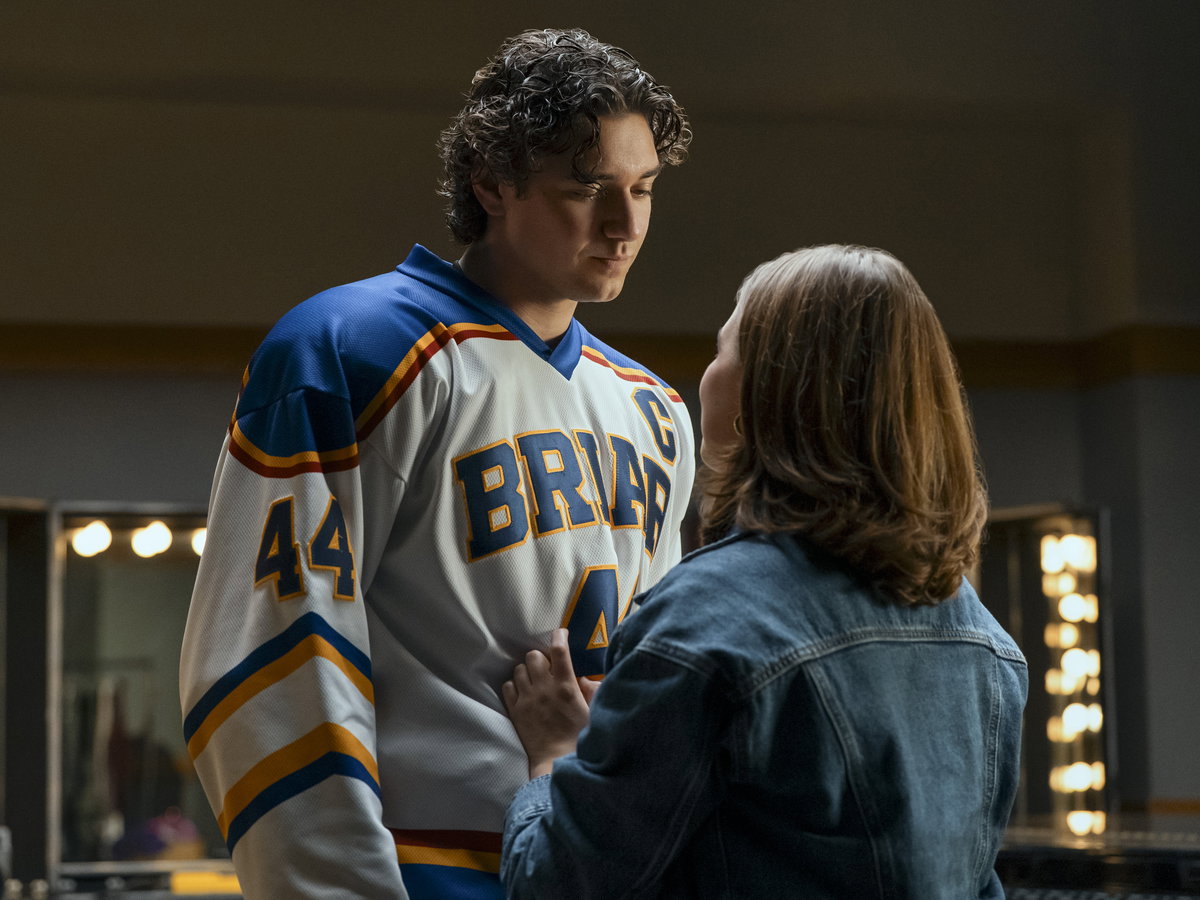 Ein junger Mann im Eishockey-Trikot steht einer jungen Frau in Jeansjacke gegenüber und guckt zu ihr herunter.