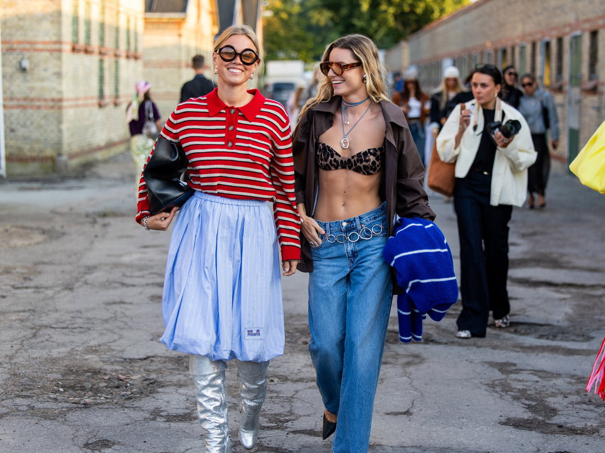 Zwei Frauen am Schlendern in der Stadt, eine mit einem Polo-Langarmshirt und einem blauen Rock, die andere mit einer Jeans und einem Leo-Bikini-Oberteil.