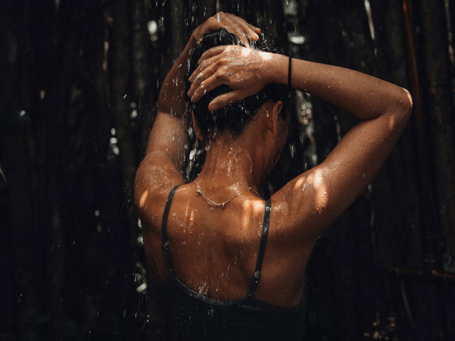 Eine Frau steht mit dem Rücken zur Kamera in einer Dusche und lässt sich mit den Armen über dem Kopf berieseln.