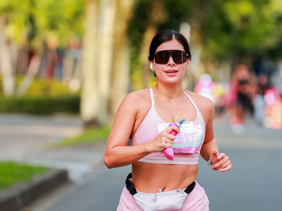 Eine junge Frau mit einer Sonnenbrille, Sportkleidung und einer kleinen Wasserflasche am Laufen auf der Straße.