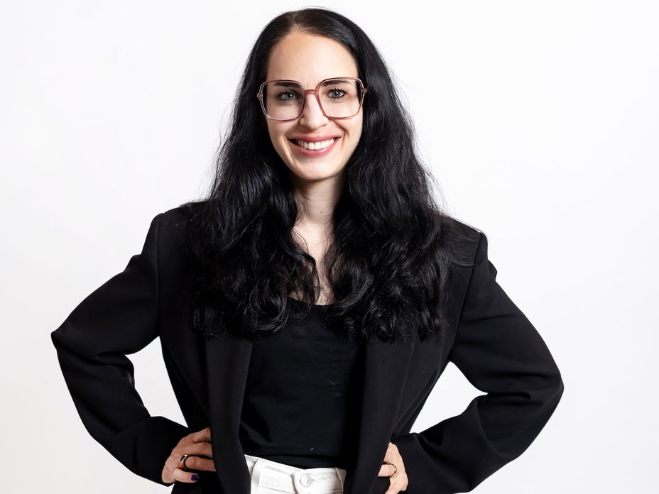 Eine Frau mit schwarzen Haaren, einem schwarzen Blazer und weißer Hose sowie einer Brille mit auf die Hüfte gestemmten Armen.