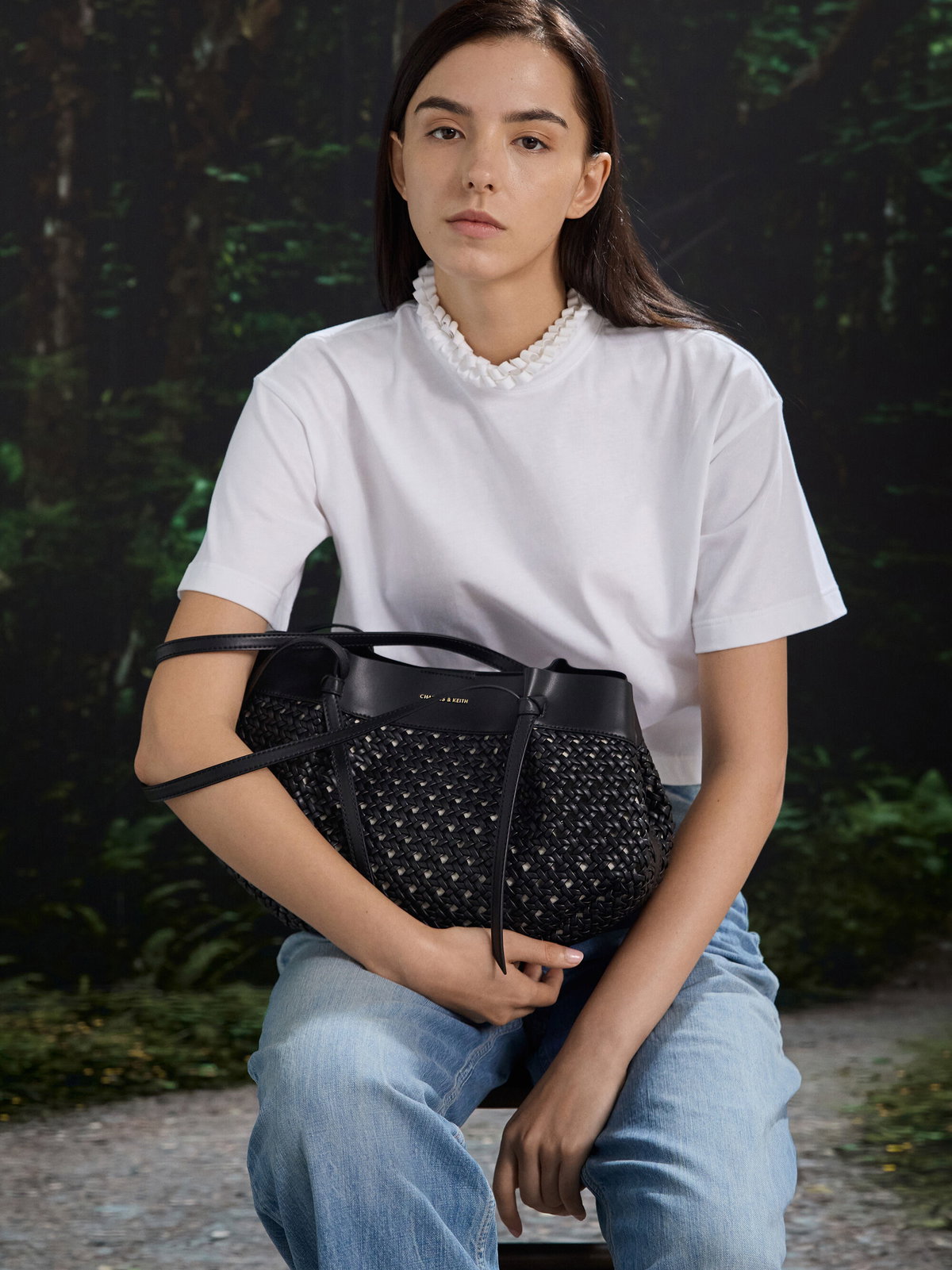 Frau mit weißem Shirt und Jeans hat eine gewebte schwarze Tasche auf dem Schoß.