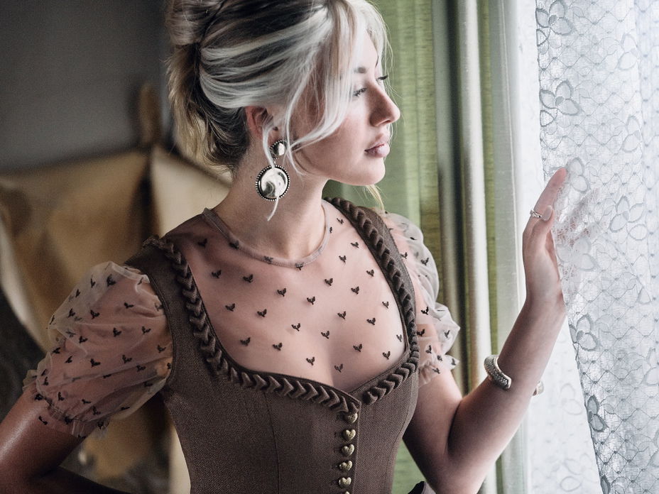 Frau mit hochgesteckten Haaren und Dirndl mit transparenter Bluse guckt durch ein Fester zur Seite.