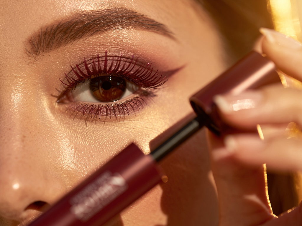 Nahaufnahme: Auge mit weinroter Mascara, offene Tube im Vordergrund, warmes Herbstlicht, echte Haut